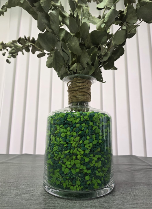 Jarrón de cristal transparente relleno de piedras decorativas combinadas en tonos verdes, con ramas de eucalipto y detalle de cuerda natural, ideal para decorar hogares, oficinas y eventos.